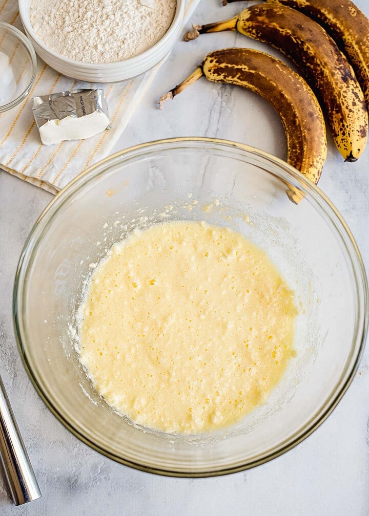 Glass bowl with beaten banana bread batter, ripe bananas, flour, and butter on a counter—perfect for making classic banana bread or adding a delicious streusel topping.