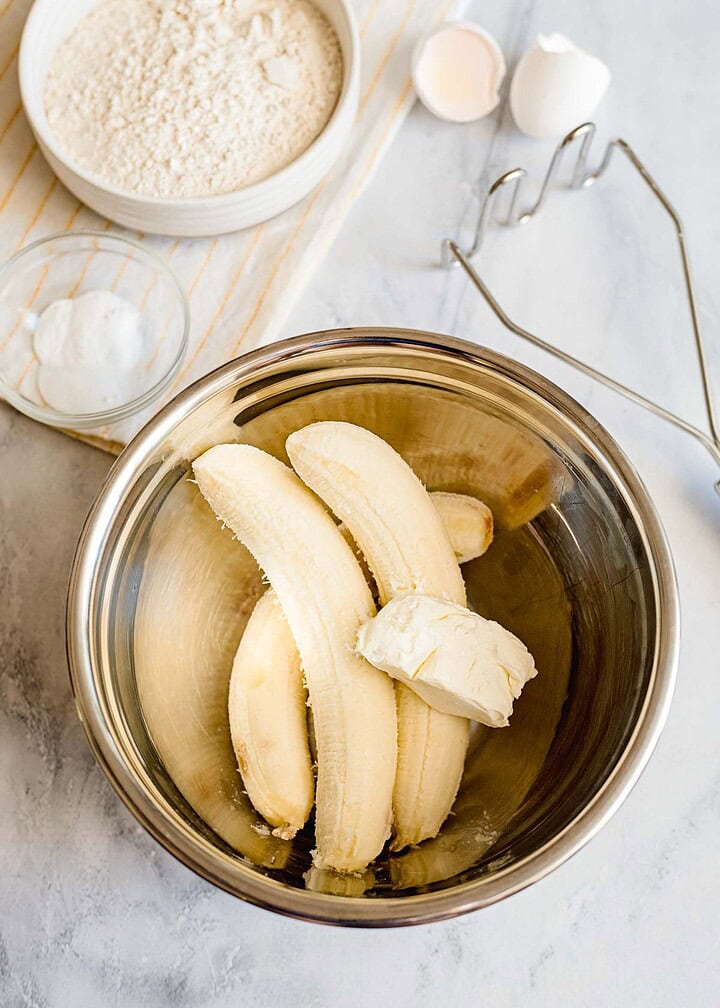 Three bananas and a chunk of butter in a metal mixing bowl, ready to be transformed into delicious banana bread, with baking ingredients and utensils nearby—perfect for adding a sweet streusel topping.