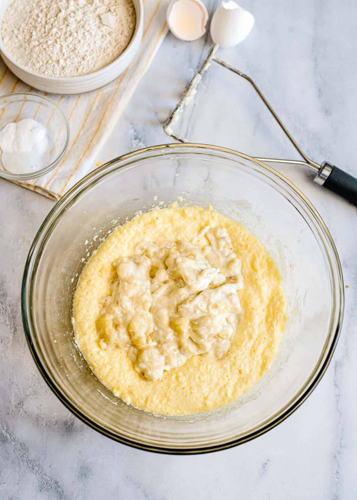 A glass bowl with mashed bananas and batter for a delicious Banana Bread Recipe, surrounded by baking ingredients on a marble surface.