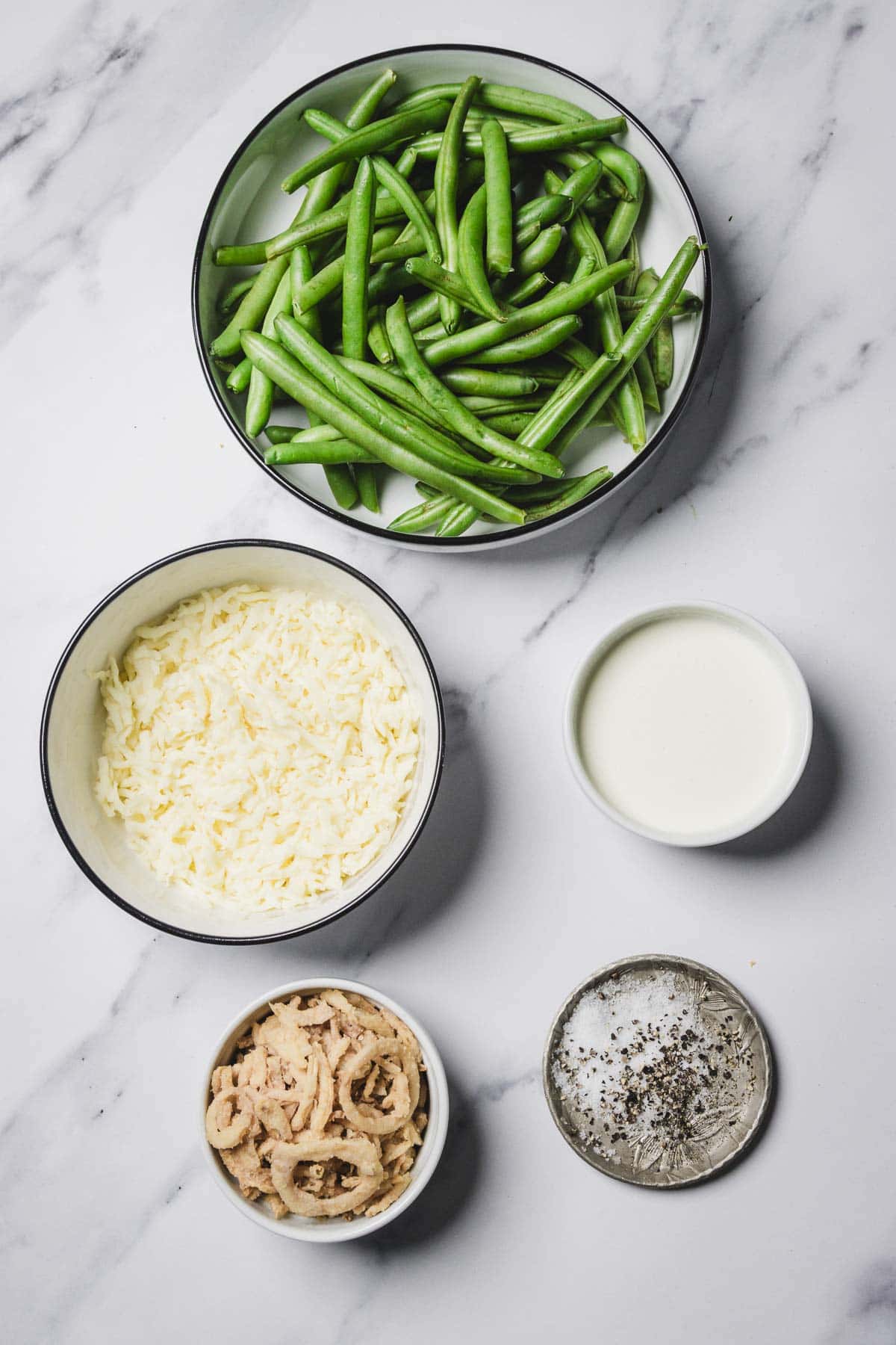 Bowls of fresh green beans, shredded cheese, cream, fried onions, and salt with pepper on a marble surface—perfect ingredients for a classic cheesy green bean casserole.