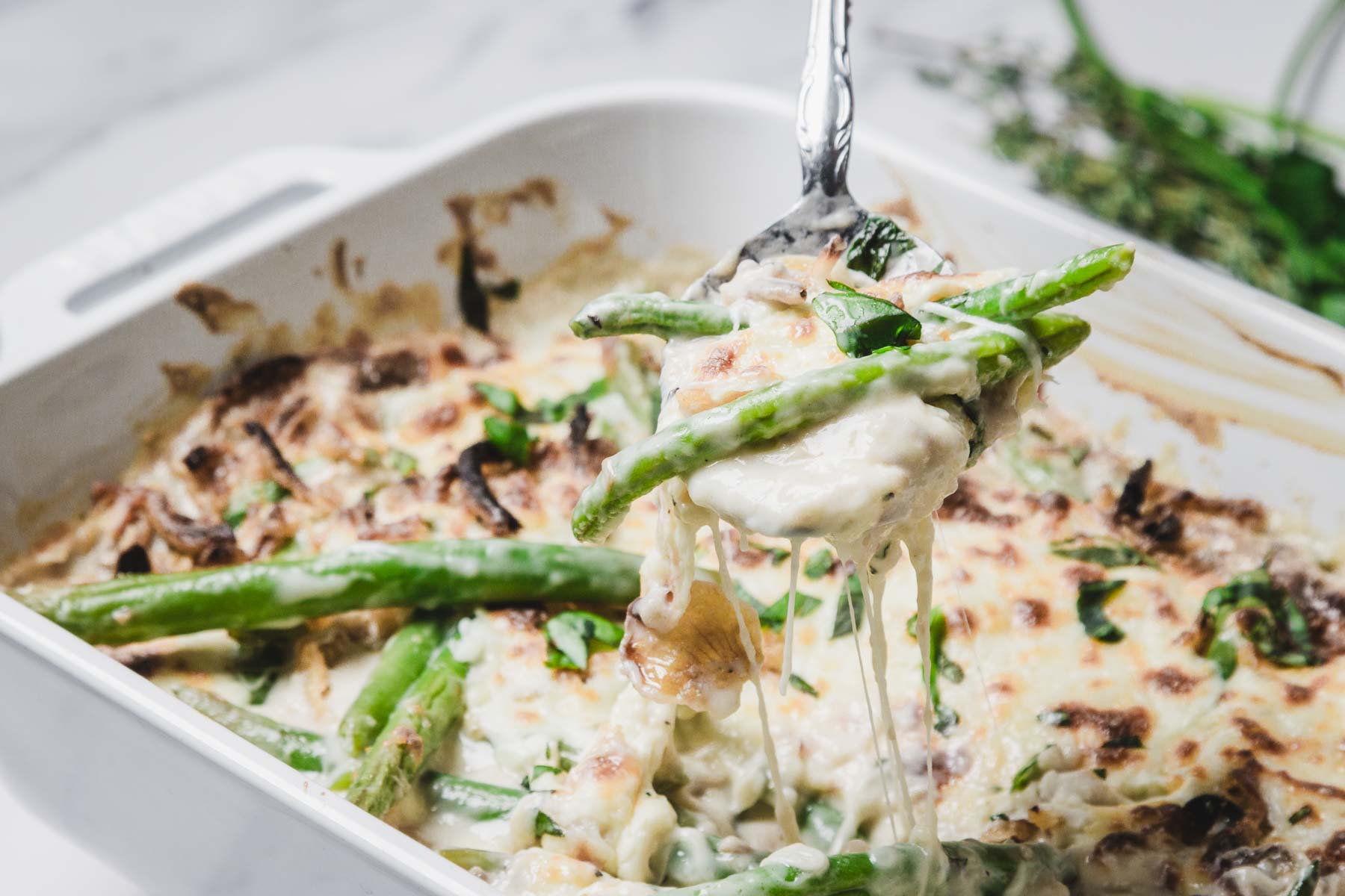 A spoon lifts a cheesy green bean casserole, made with fresh green beans, from a white baking dish and garnished with herbs.