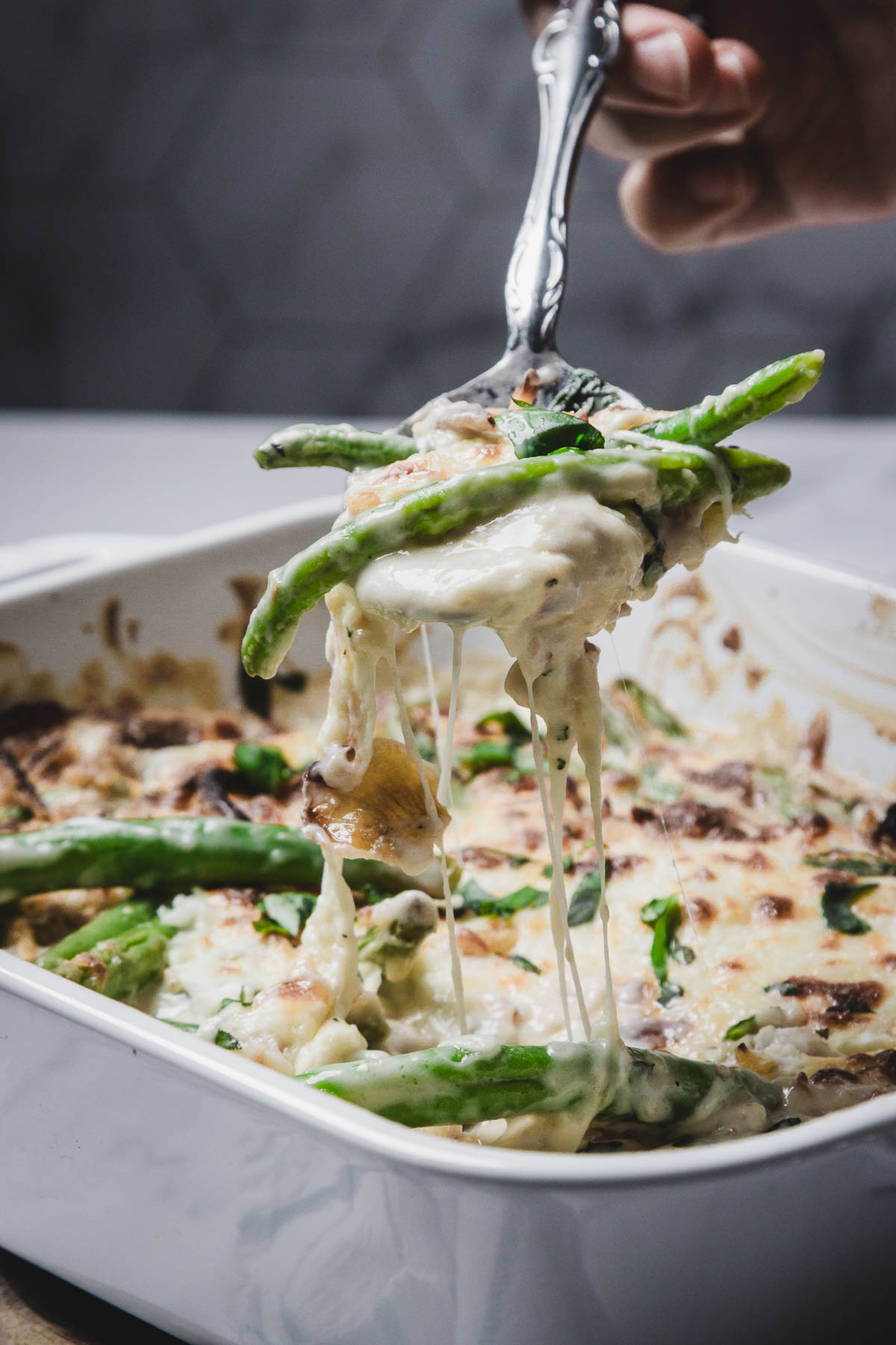 A fork lifts Fresh Green Beans from a cheesy green bean casserole in a white baking dish, revealing gooey melted cheese strands.