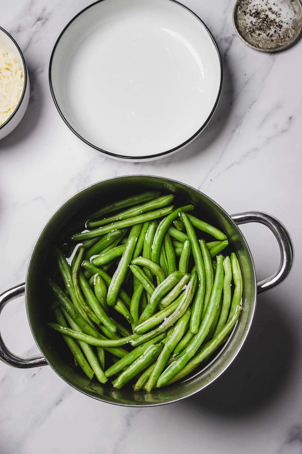 A pot of fresh green beans in water sits on a marble countertop beside an empty plate, perfect for making a delicious cheesy green bean casserole.