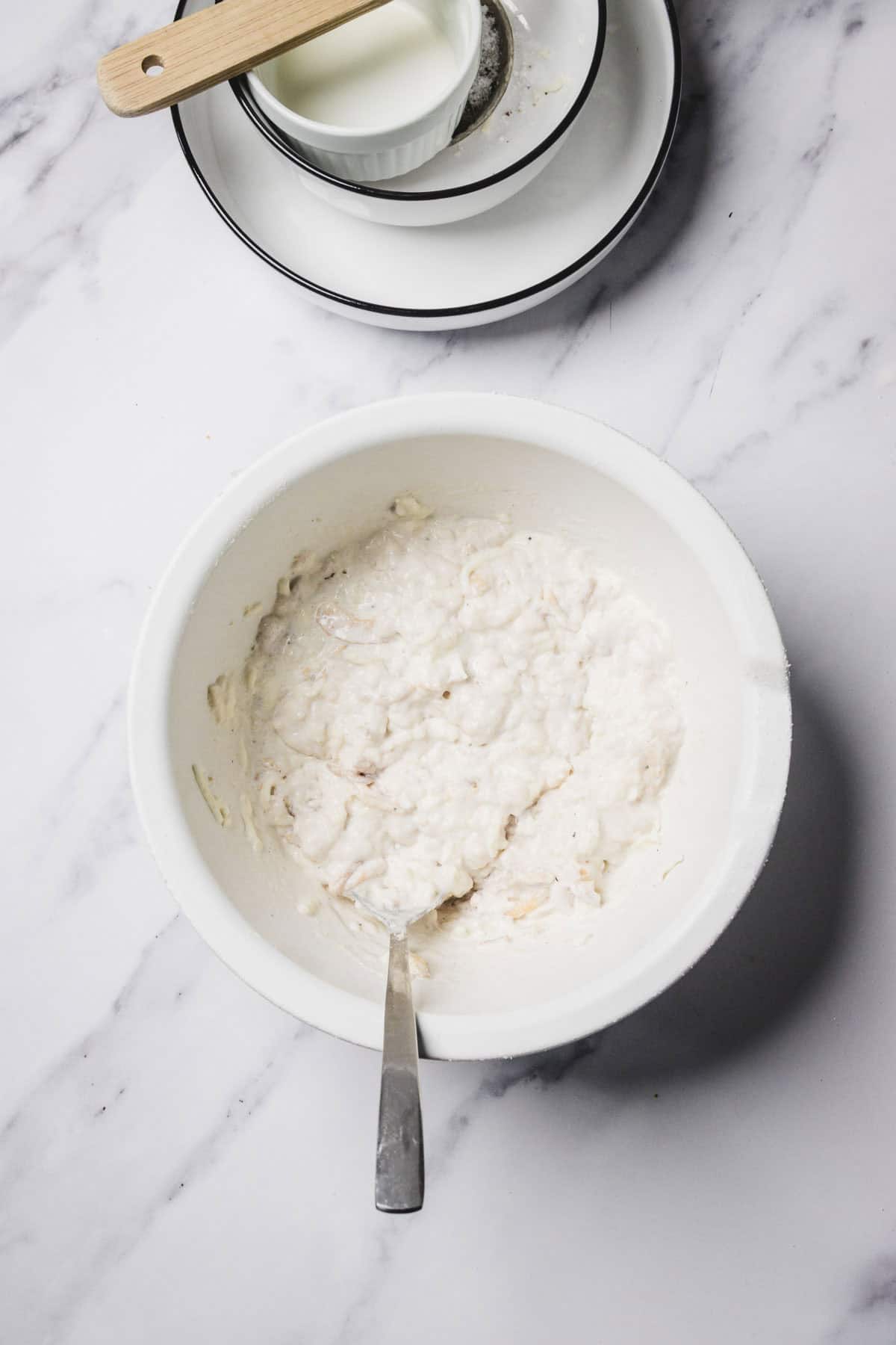 White bowl with lumpy batter and a spoon, next to stacked plates and a measuring cup of milk on a marble surface—perfect prep for making cheesy green bean casserole with fresh green beans.