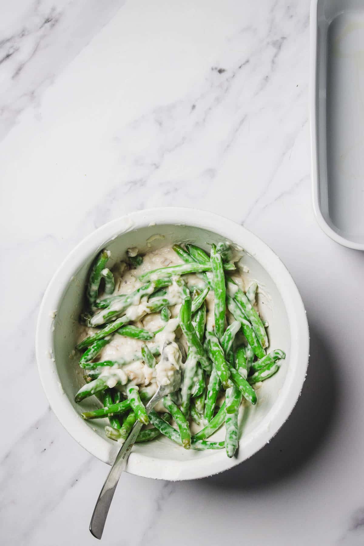 A bowl of cheesy green bean casserole in creamy sauce with a spoon, set on a marble surface.