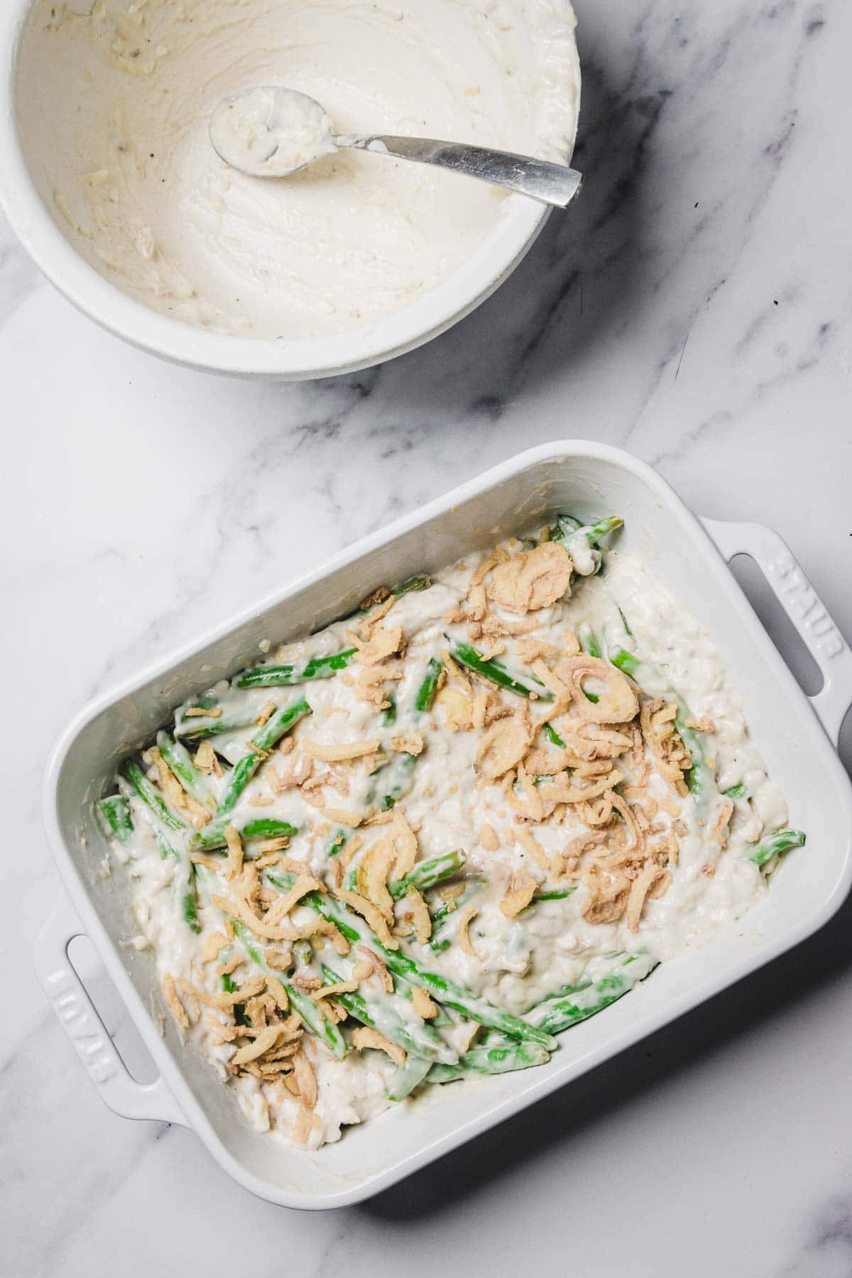 A baking dish of Cheesy Green Bean Casserole topped with fried onions sits beside a mixing bowl with a spoon.