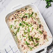 Baked cheesy green bean casserole topped with melted cheese and fresh herbs in a white dish on a checked towel.