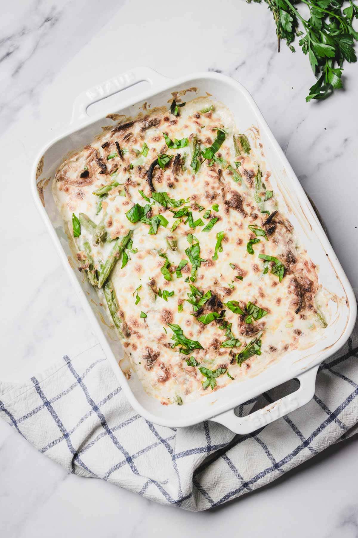 Baked cheesy green bean casserole topped with melted cheese and fresh herbs in a white dish on a checked towel.