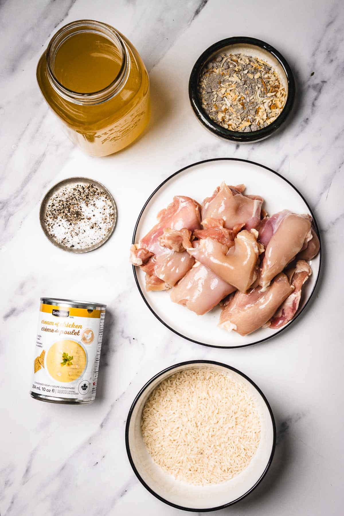 Raw chicken, uncooked rice, seasonings, chicken broth, and a can of cream soup arranged on a marble surface.