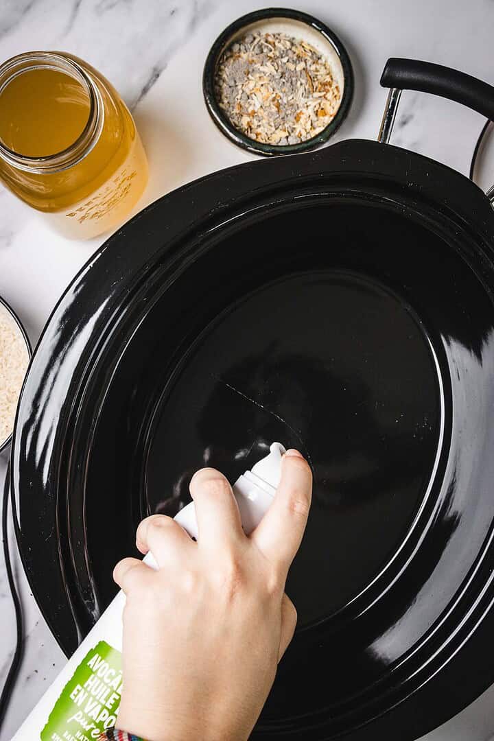 Hand spraying non-stick oil into a black slow cooker with seasonings and broth nearby on a marble counter, prepping for delicious crockpot recipes like chicken and rice.