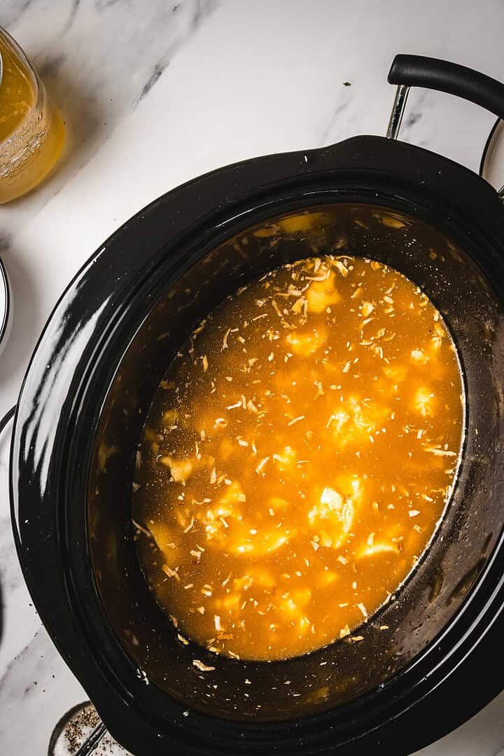A Crockpot chicken dish with shredded meat and savory broth sits on a white marble countertop—a cozy favorite for easy crockpot recipes.