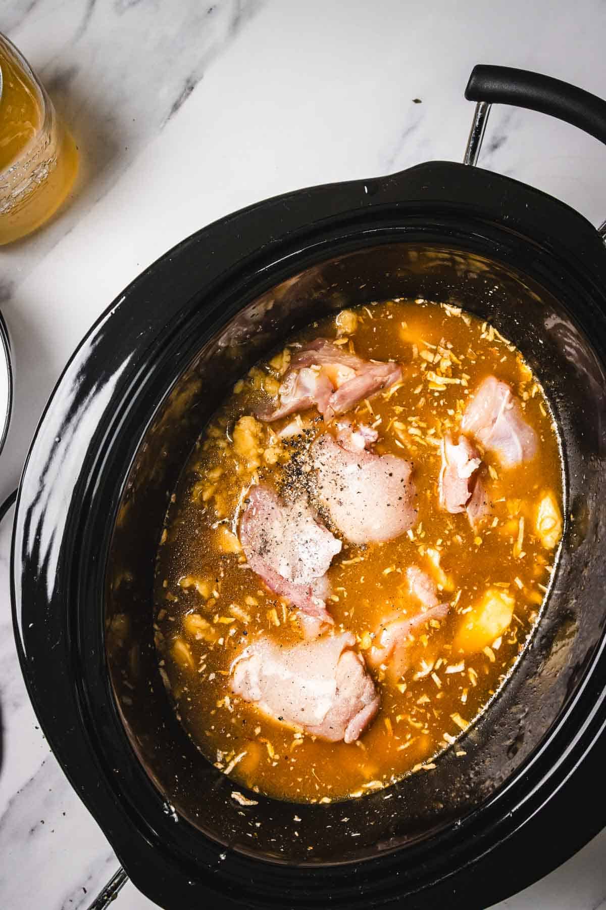 Raw chicken thighs with broth, seasonings, and garlic in a black Crockpot for an easy crockpot chicken recipe on a white marble counter.