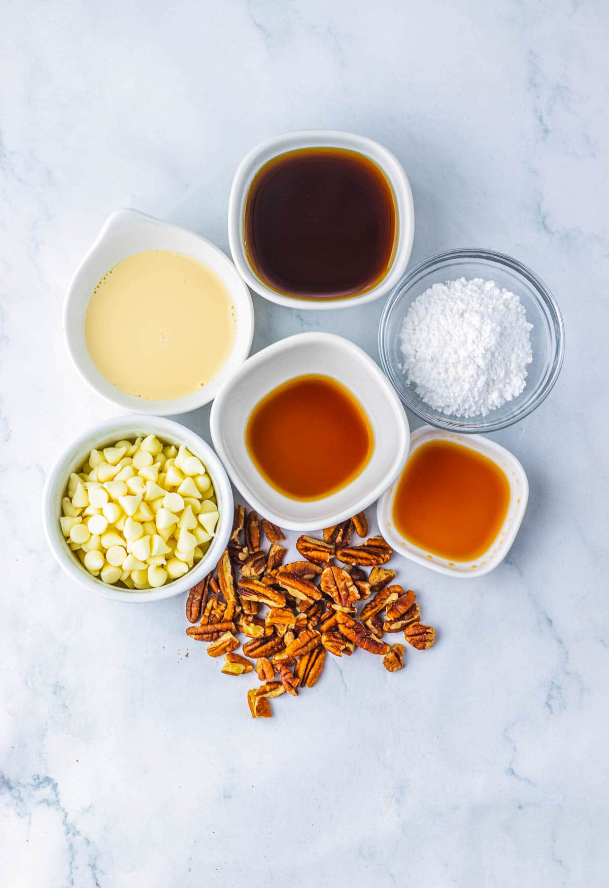 Assorted bowls of white chocolate chips, pecans, syrup, condensed milk, vanilla, and powdered sugar on a marble surface&mdash;perfect for crafting a delicious Pecan Pie Fudge recipe.