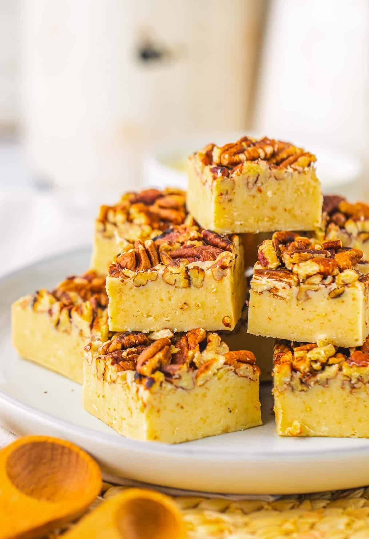 Squares of pecan pie fudge are stacked on a white plate, with wooden spoons in the foreground&mdash;a tempting treat for anyone seeking a rich pecan pie fudge recipe.