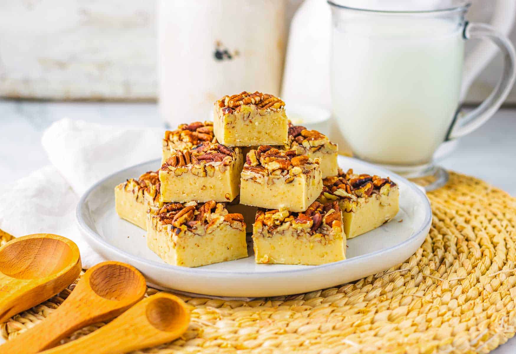 A plate of Pecan Pie Fudge squares topped with pecans sits next to a glass of milk on a woven mat, with wooden spoons nearby&mdash;perfect for those searching for the ultimate Fudge Recipe.