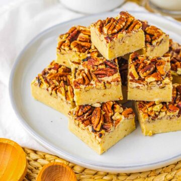 Pecan Pie Fudge Recipe. A plate of pecan-topped fudge squares stacked neatly, with baking tools and ingredients in the background.