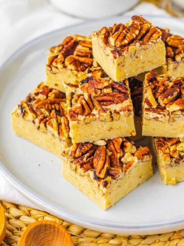 Pecan Pie Fudge Recipe. A plate of pecan-topped fudge squares stacked neatly, with baking tools and ingredients in the background.