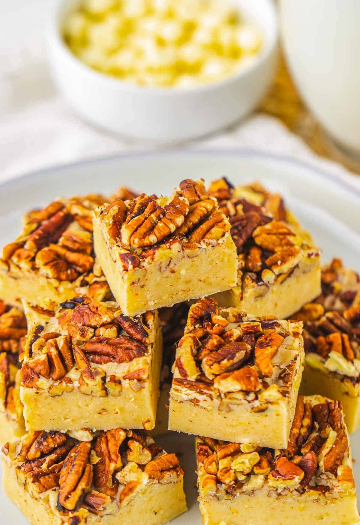 Squares of creamy pecan pie fudge topped with chopped pecans, stacked on a white plate.