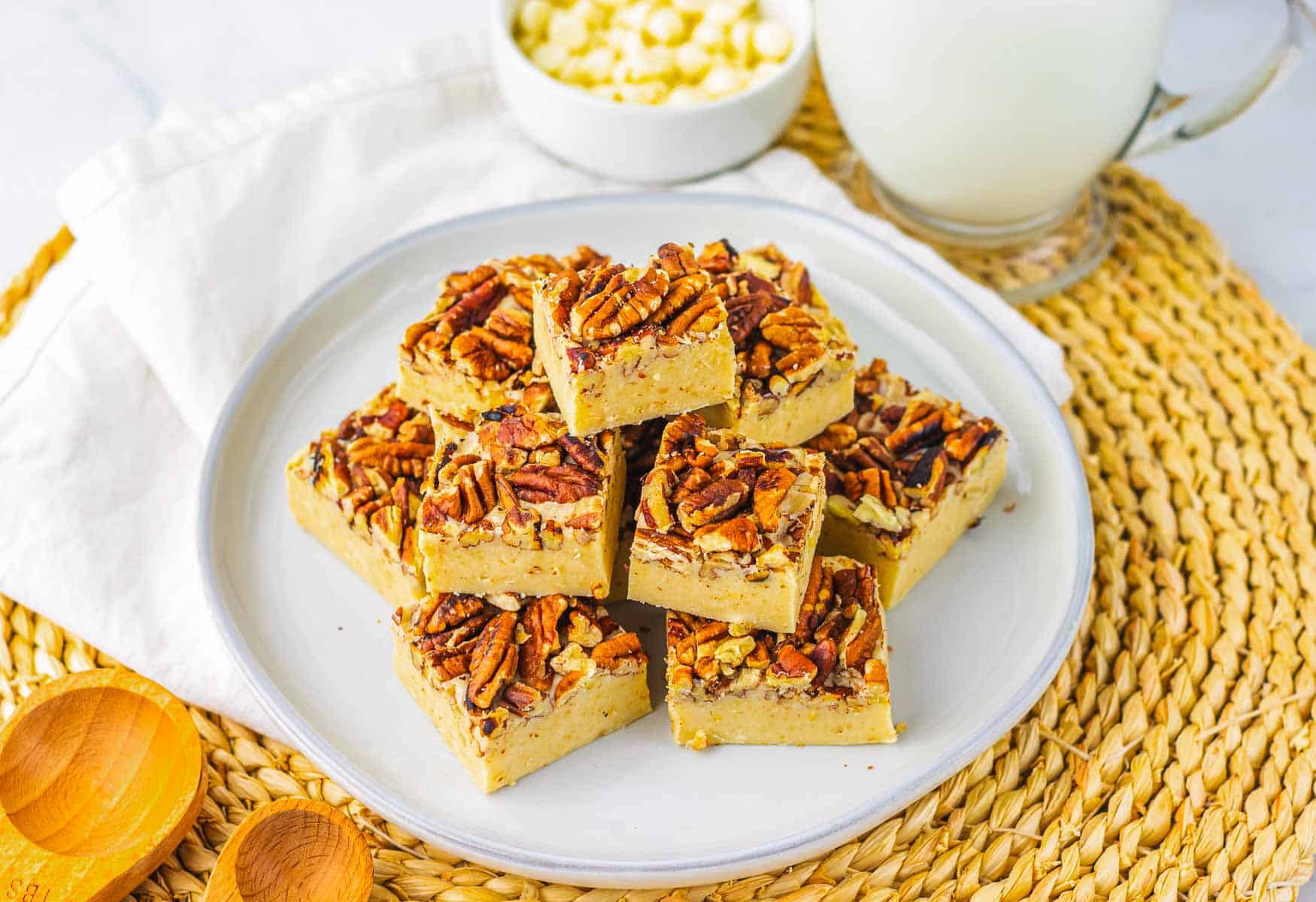 Nine pieces of pecan pie fudge stacked on a white plate, with a bowl of white chips and a glass of milk nearby&mdash;a delicious twist on the classic pecan pie recipe.