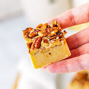 Pecan Pie Fudge Recipe. A hand holding a square dessert topped with pecans and other nuts, this treat looks just like Pecan Pie Fudge—perfect for anyone searching for an irresistible fudge recipe.