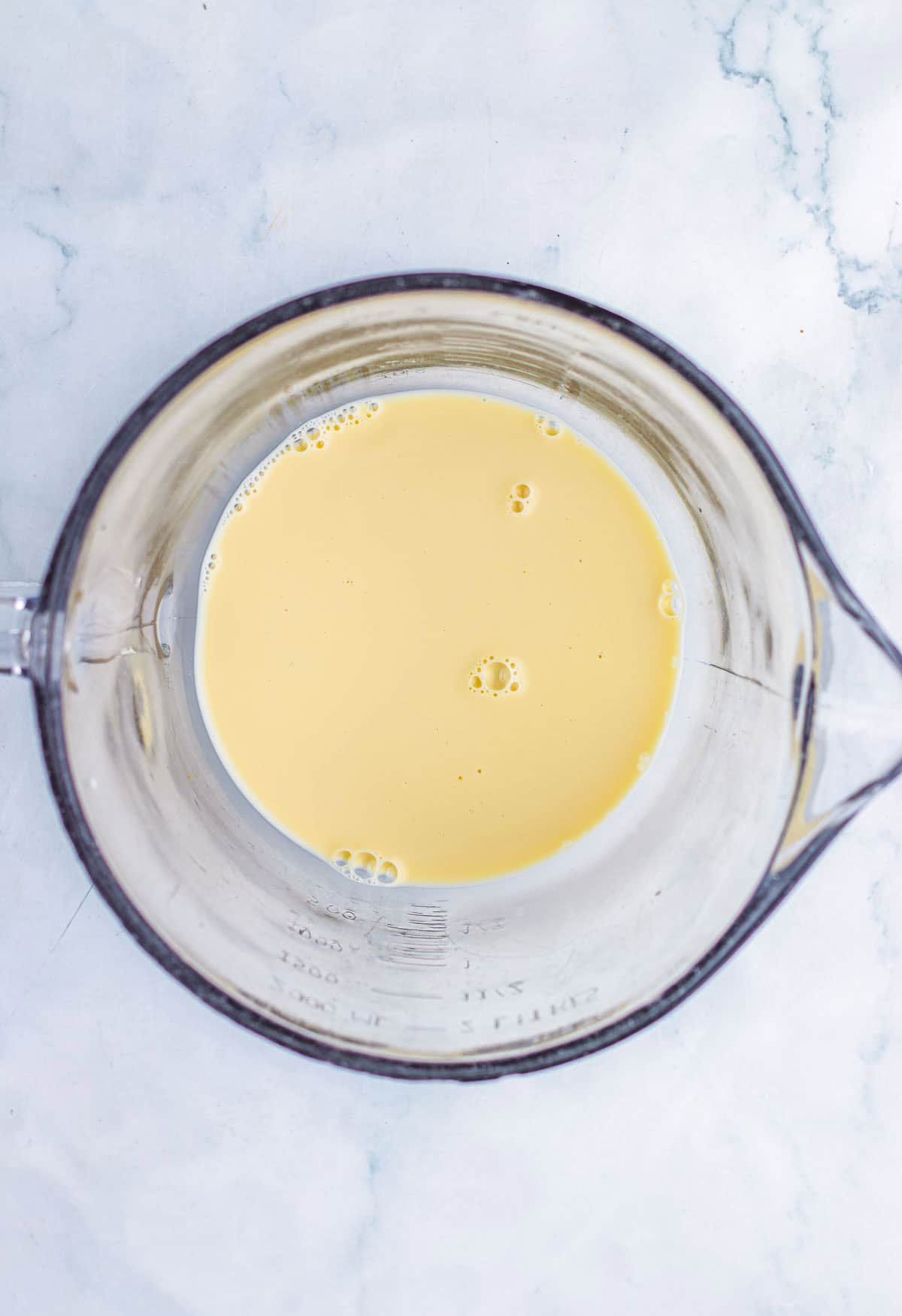 Top view of a glass measuring cup filled with a light yellow liquid on a white marble surface, ready for making the perfect Pecan Pie Fudge Recipe.
