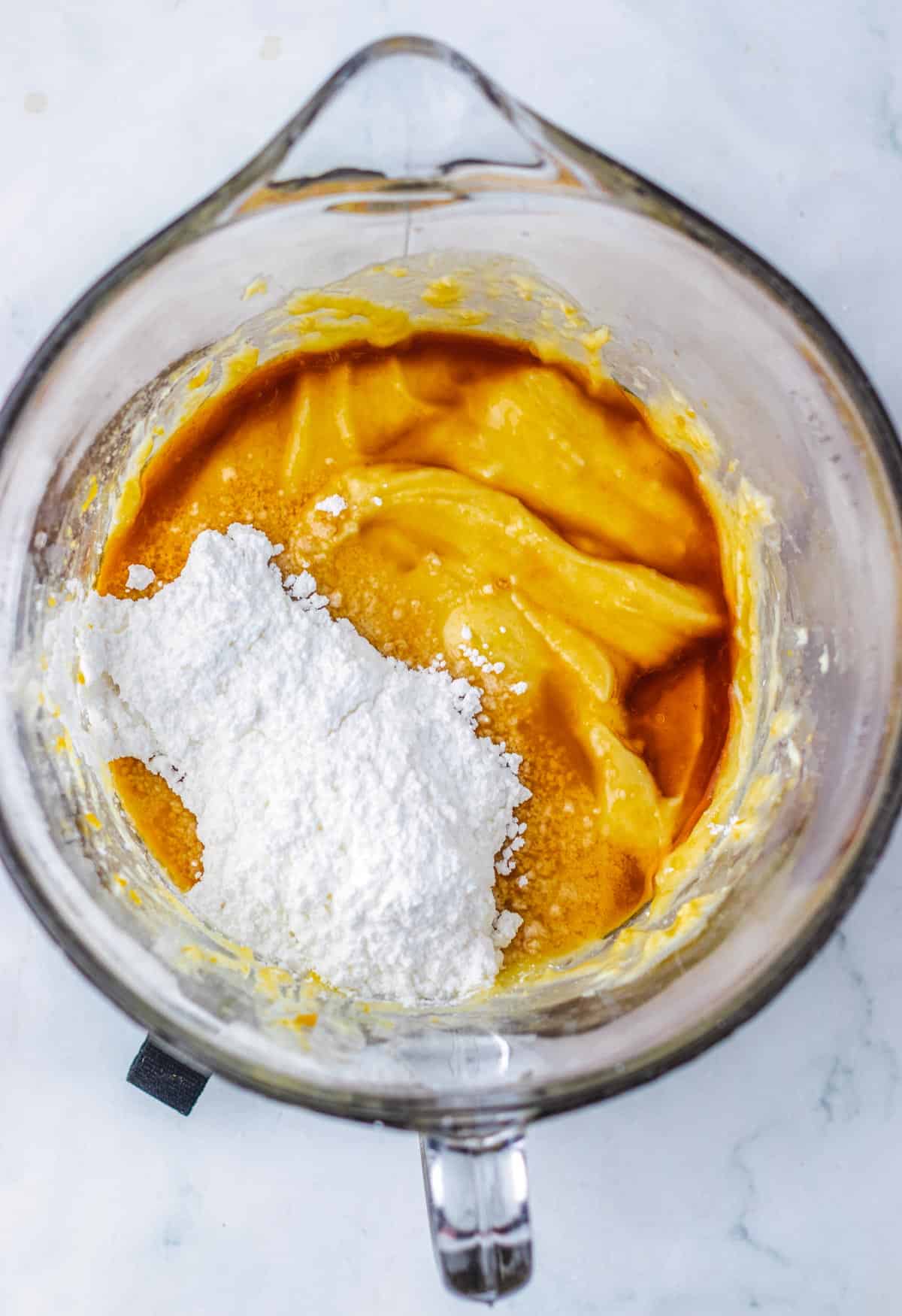 Glass mixing bowl with yellow batter and a pile of white powdered sugar on top, seen from above&mdash;perfect for starting your pecan pie fudge recipe.