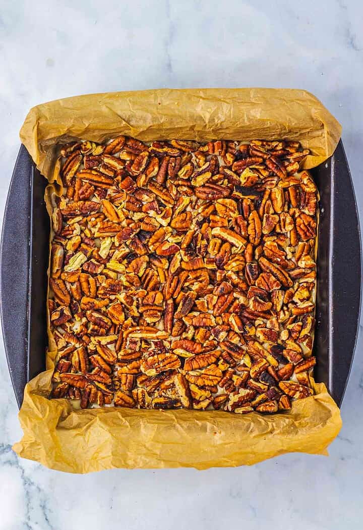 A square pan lined with parchment paper, filled with baked pecan bars that taste just like Pecan Pie Fudge, sits on a marble surface.