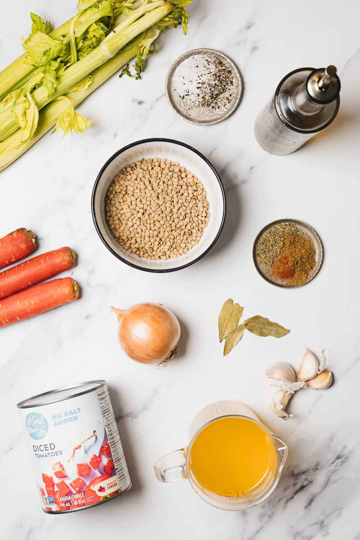 Lentils, carrots, celery, onion, garlic, spices, broth, oil, and diced tomatoes—everything you need for a hearty Slow Cooker Lentil Soup is displayed on a white marble surface.