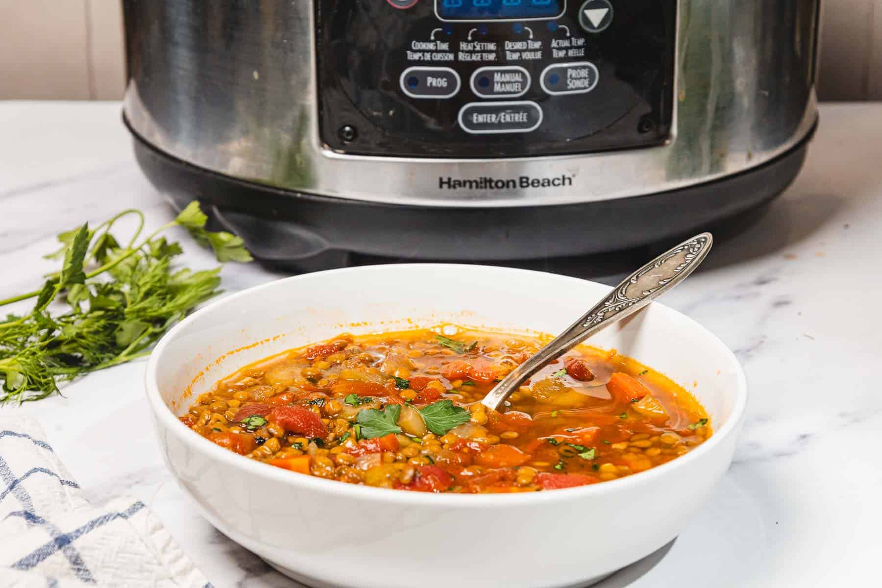 A bowl of carrot soup with a spoon sits in front of a slow cooker on a marble counter.
