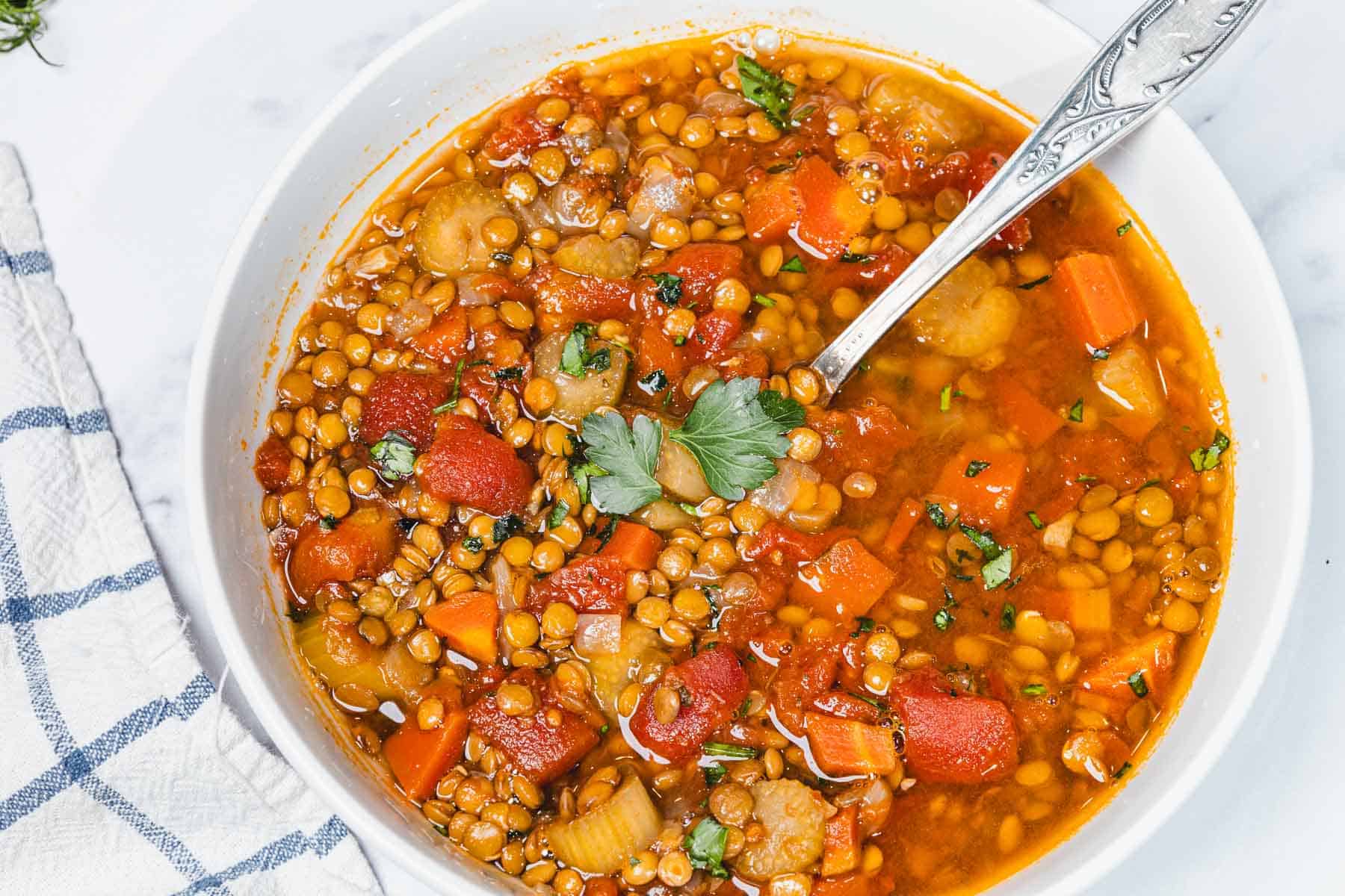 Bowl of Slow Cooker Lentil Soup with carrots, tomatoes, herbs, and a spoon, on a white surface with a checked napkin.