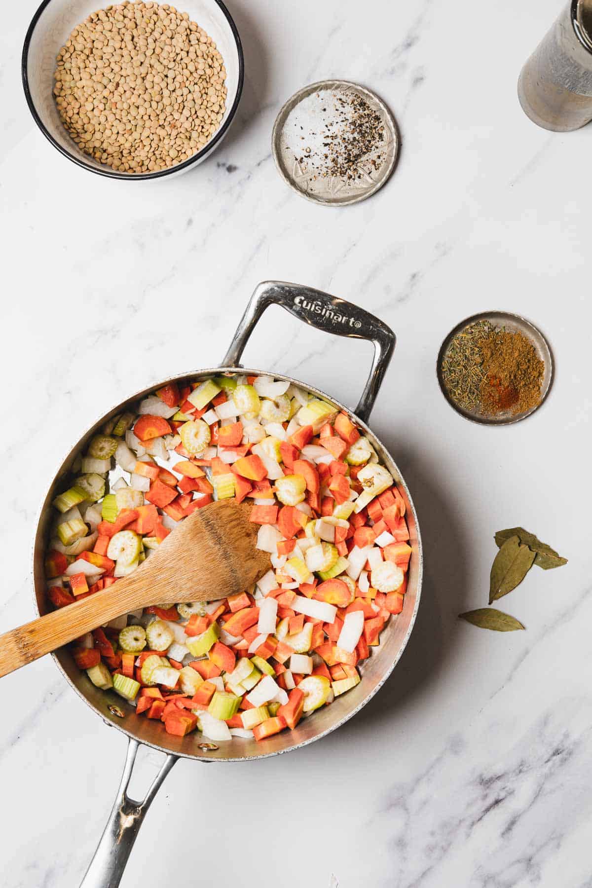 Chopped carrots, celery, and onions in a pan with a wooden spoon, surrounded by spices and lentils—the perfect base for hearty Lentil Soup.