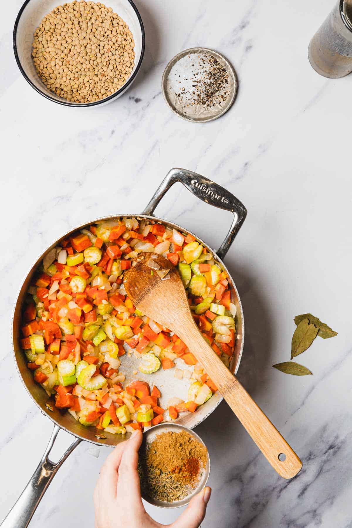 A hand adds spices to a pan of chopped vegetables for lentil soup cooking on a marble countertop.