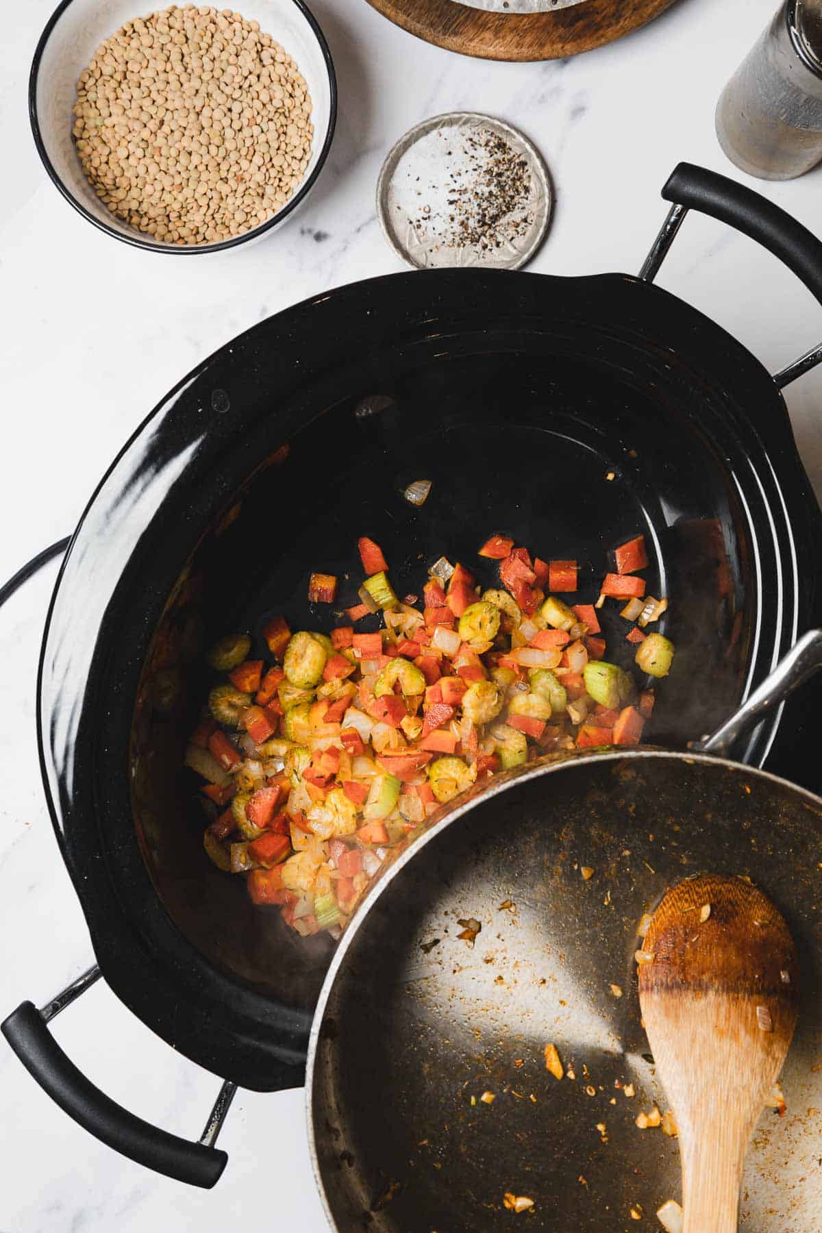 A slow cooker with sautéed vegetables, a pan and wooden spoon, plus bowls of lentils and pepper nearby—perfect for preparing hearty Lentil Soup.