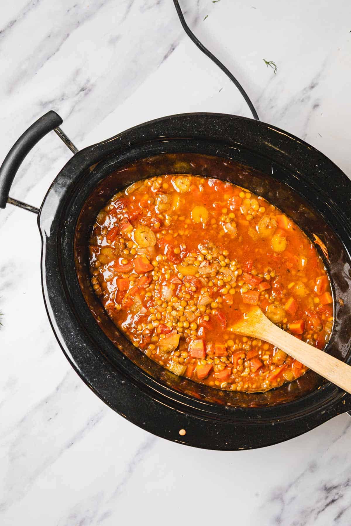 Lentil soup with vegetables simmering in a black slow cooker, being gently stirred with a wooden spoon.