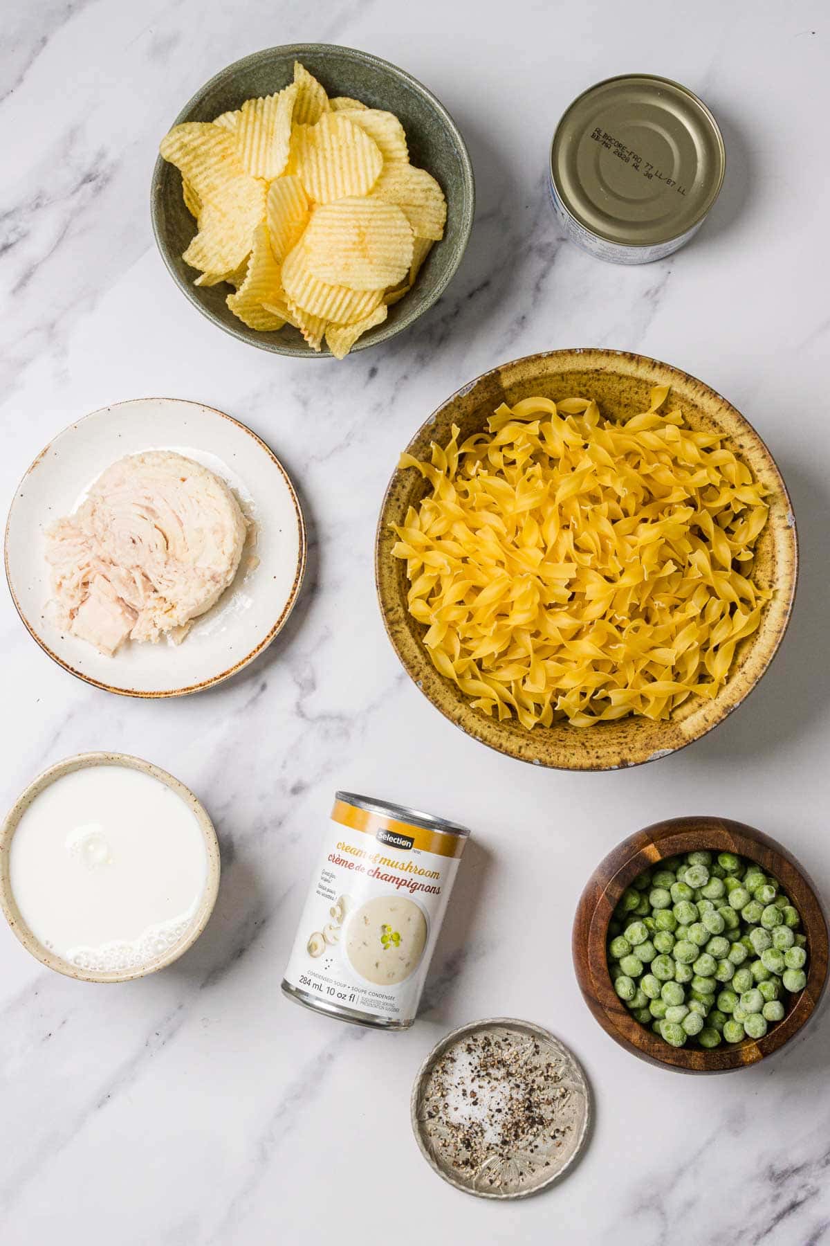 Bowls of egg noodles, potato chips, canned tuna, cream, peas, soup, and pepper on a marble surface.