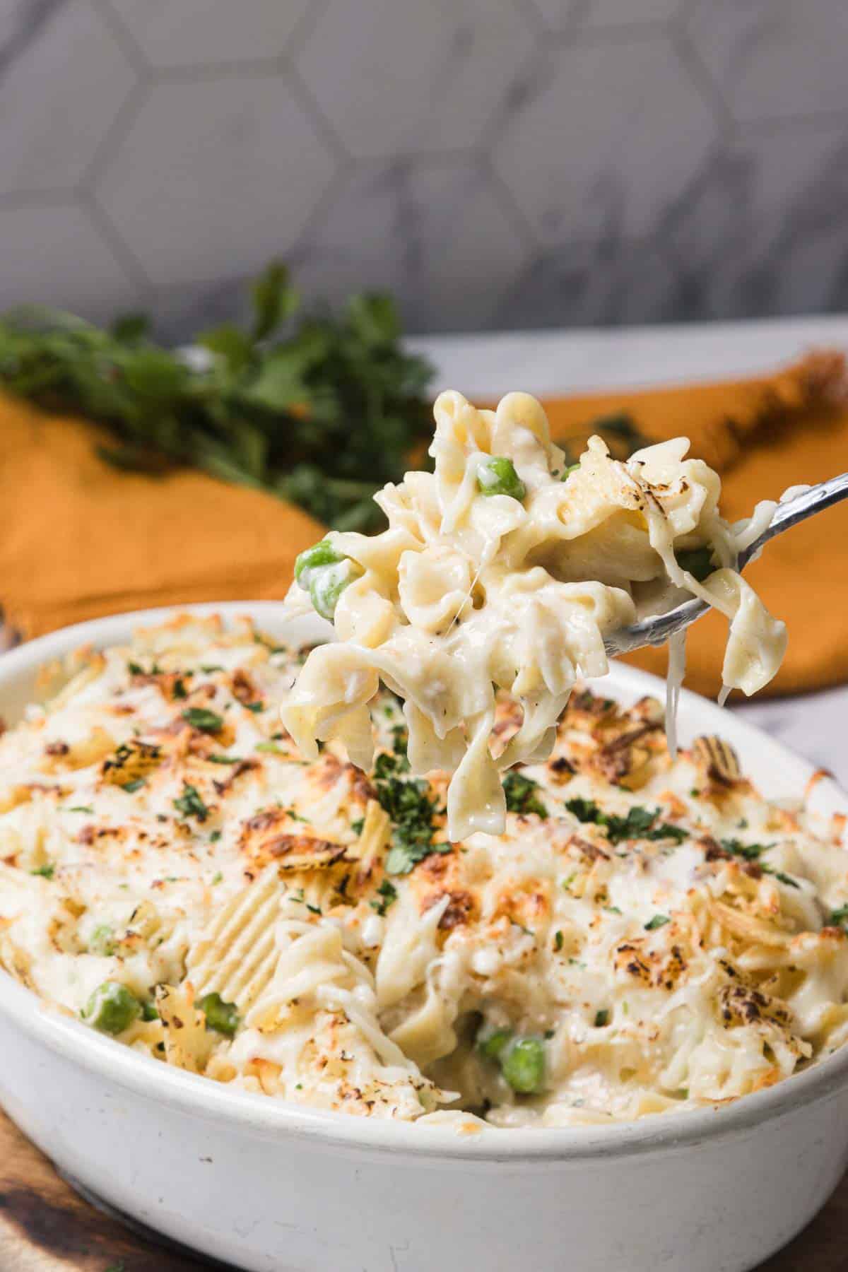 A fork lifts a cheesy tuna noodle casserole with peas from a white dish, topped with melted cheese and herbs.