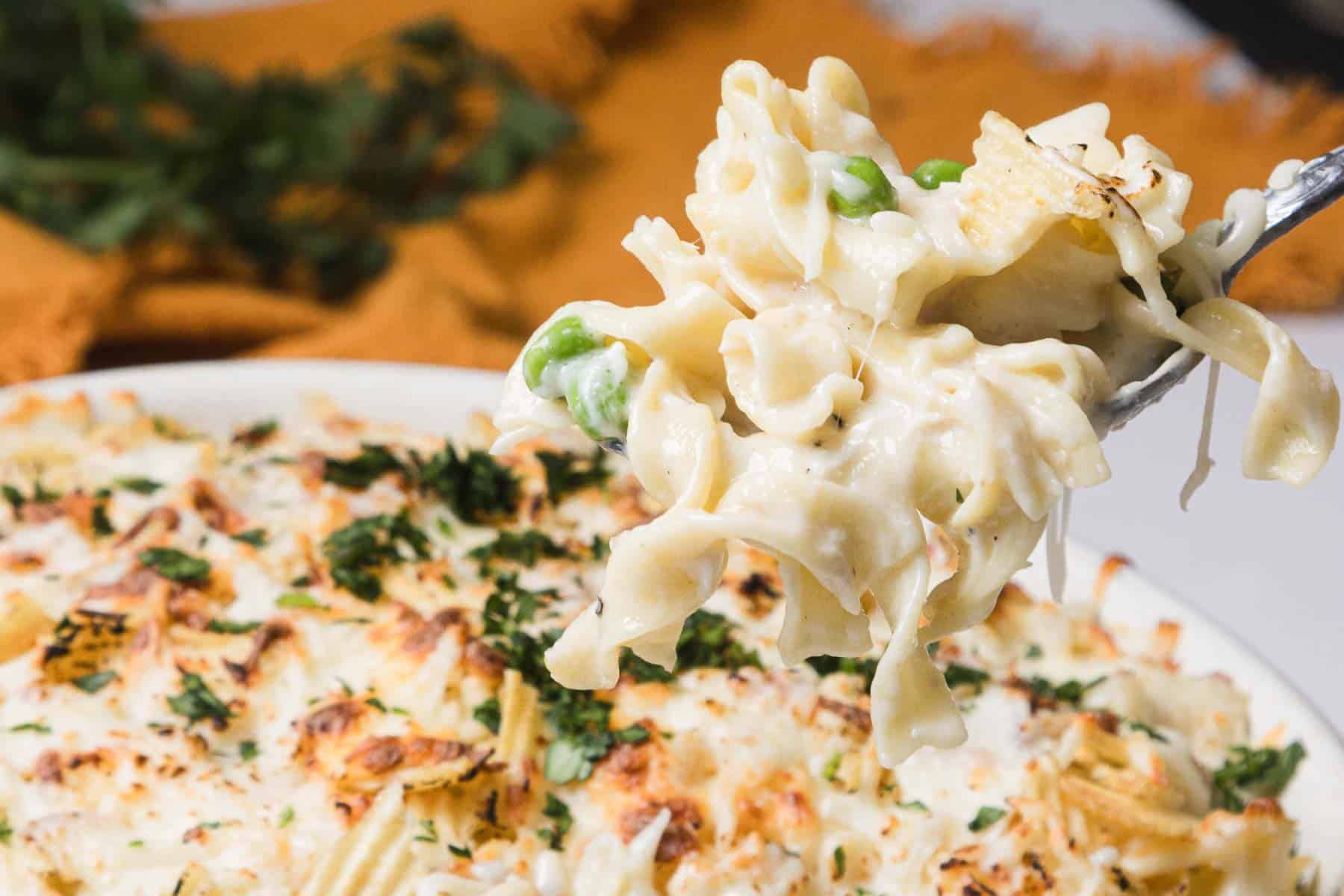 Close-up of a fork lifting creamy baked pasta with peas and herbs from a dish in the background.