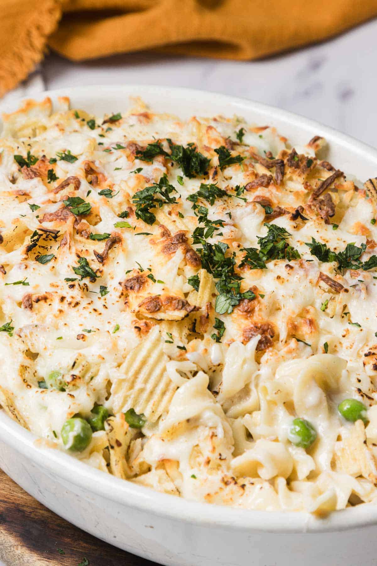 Baked casserole topped with melted cheese, herbs, and potato chips, with visible peas and pasta in a white dish.