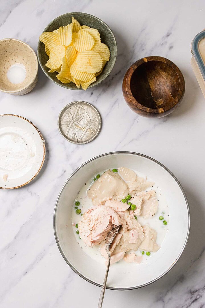 Bowl of creamy chicken and peas with chips, salt, and wooden bowl on a white marble surface, inspired by classic Tuna Noodle Casserole flavors for a comforting twist on a traditional casserole recipe.