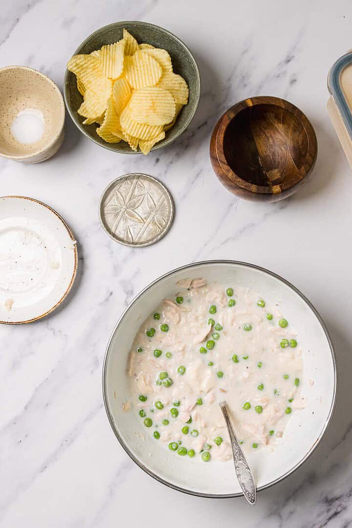 Bowl of creamy tuna and pea mixture with a spoon, surrounded by chips and small bowls on a marble surface—perfect for enjoying your favorite Tuna Casserole or as a tasty dip inspired by the classic casserole recipe.