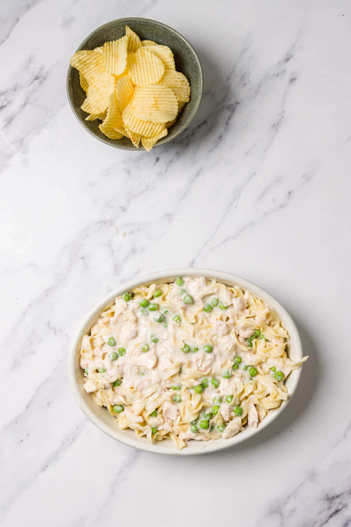 Oval dish of creamy tuna noodle casserole with peas, next to a bowl of ridged potato chips on a white surface. Perfect comfort food recipe for any gathering.