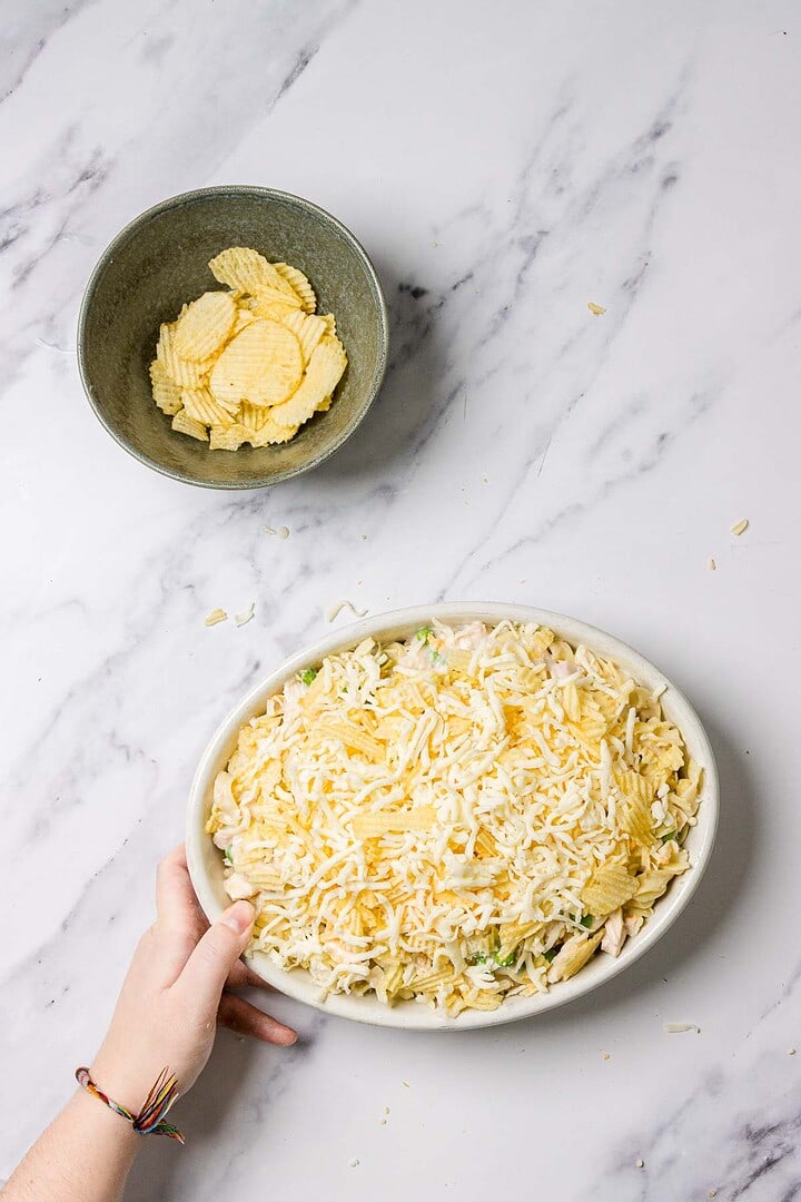 A hand holds a Tuna Noodle Casserole dish topped with shredded cheese, next to a bowl of ridged potato chips on a marble surface.