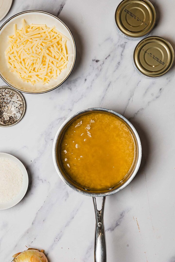 A saucepan of bubbling broth on a marble counter with shredded cheese, cans of tuna, onions, and seasonings nearby&mdash;perfect for preparing a hearty Tuna Casserole.