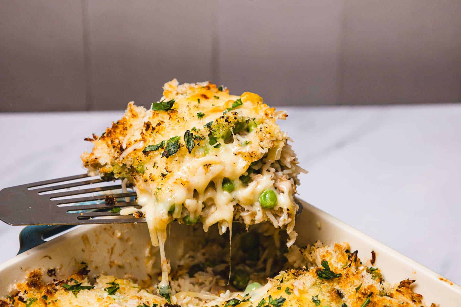 A cheesy Tuna Rice Casserole with peas and herbs is served hot from a baking dish with a spatula, perfect for a comforting family meal.