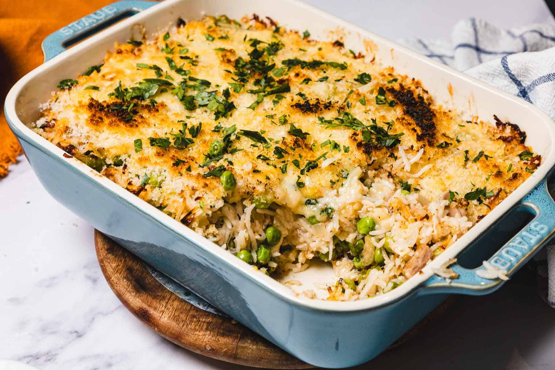 A casserole dish with baked rice, peas, melted cheese, herbs, and tuna, with a serving scooped out&mdash;classic tuna casserole comfort in every bite.