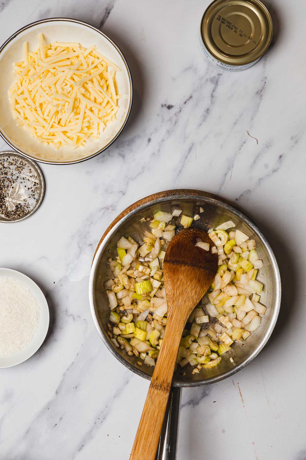 A pan with chopped onions and leeks being saut&eacute;ed, surrounded by cheese, pepper, and a can of tuna on a marble surface&mdash;perfect ingredients for a delicious Tuna Casserole.