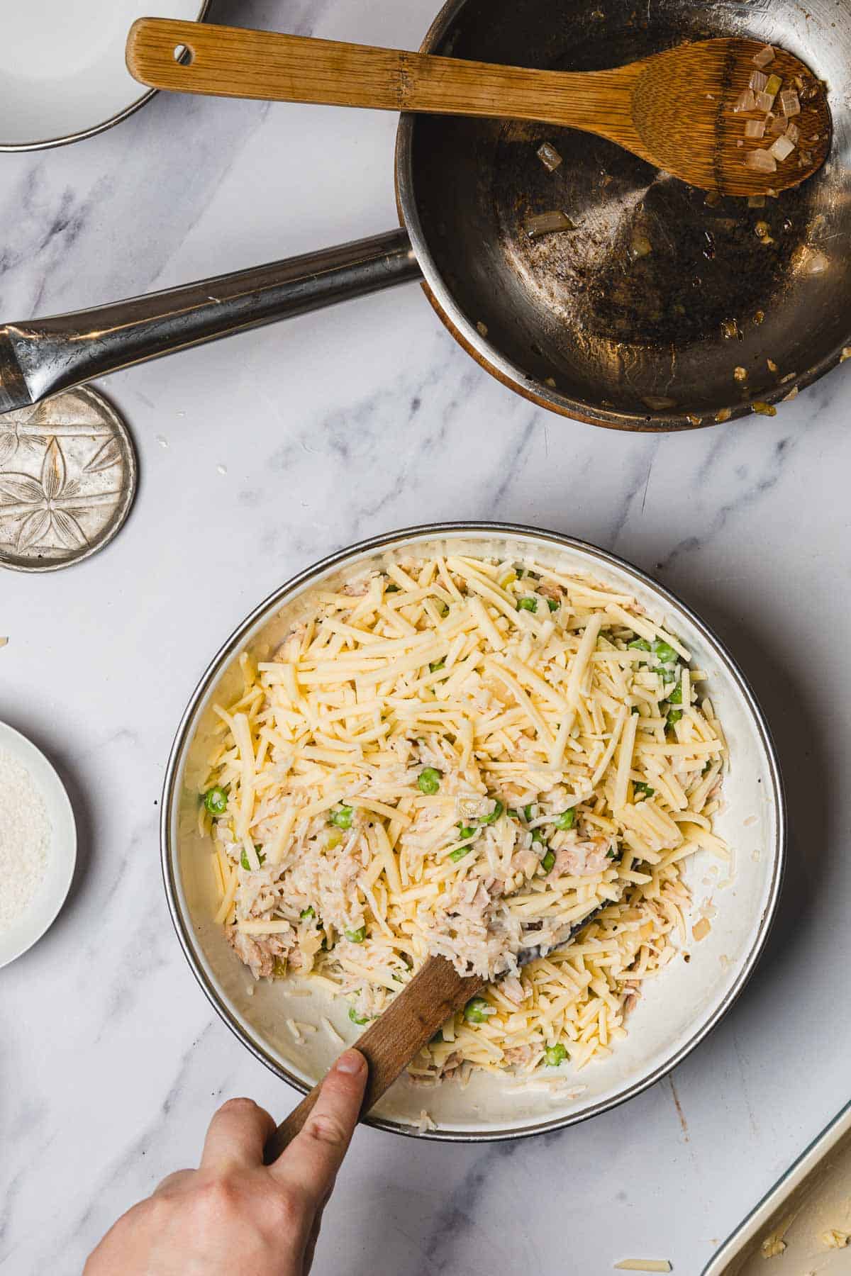 A hand mixing shredded cheese, rice, herbs, and tuna in a bowl next to a skillet with saut&eacute;ed onions&mdash;perfect for assembling a flavorful Tuna Casserole.