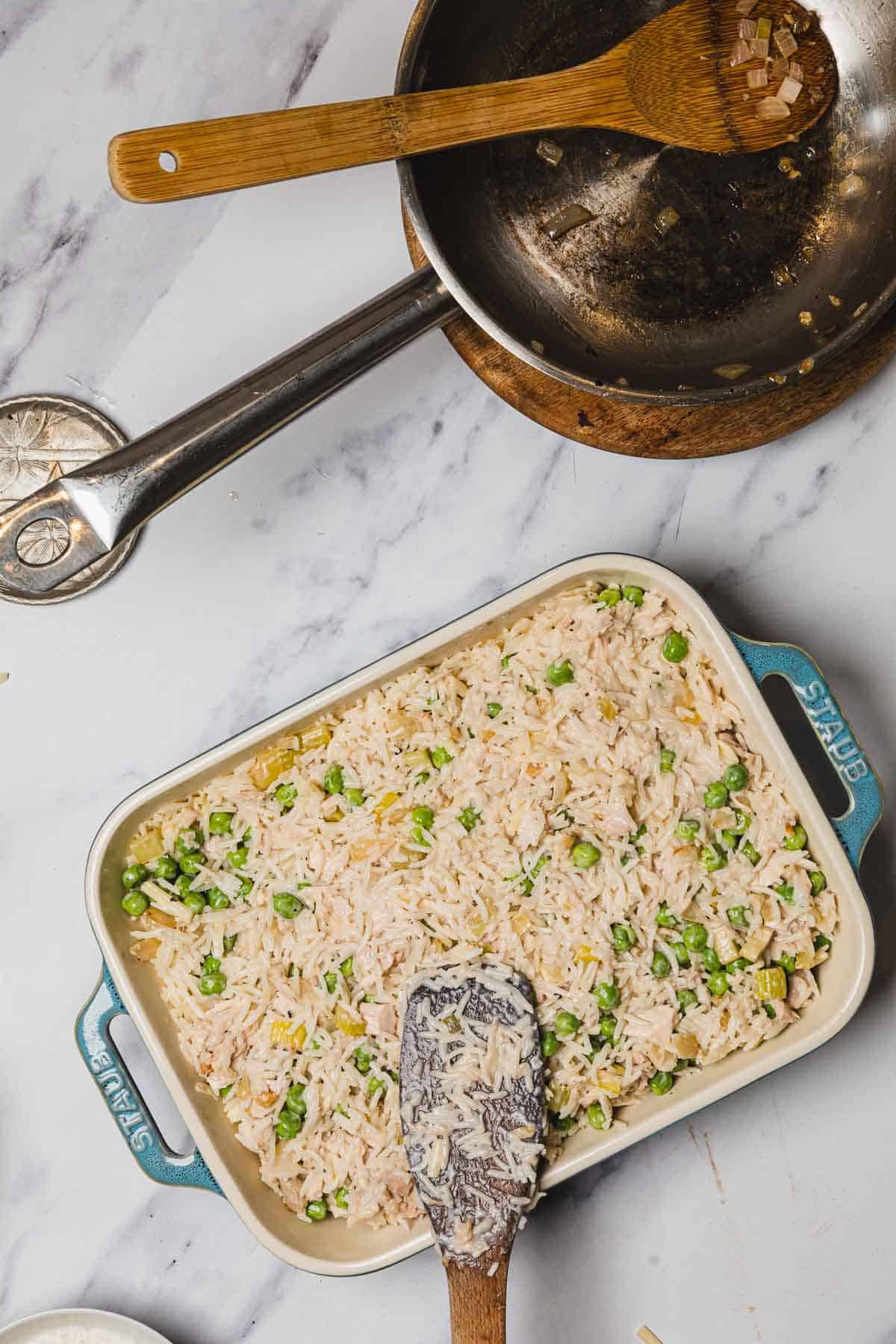 A baking dish with creamy rice and peas casserole sits beside a pan and wooden spoon on a marble countertop.