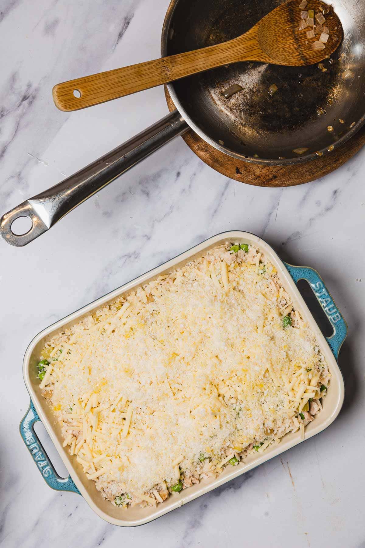 Rectangular baking dish with shredded cheese on top; rice casserole with tuna, wooden spoon, and pan with onions beside it on marble surface.