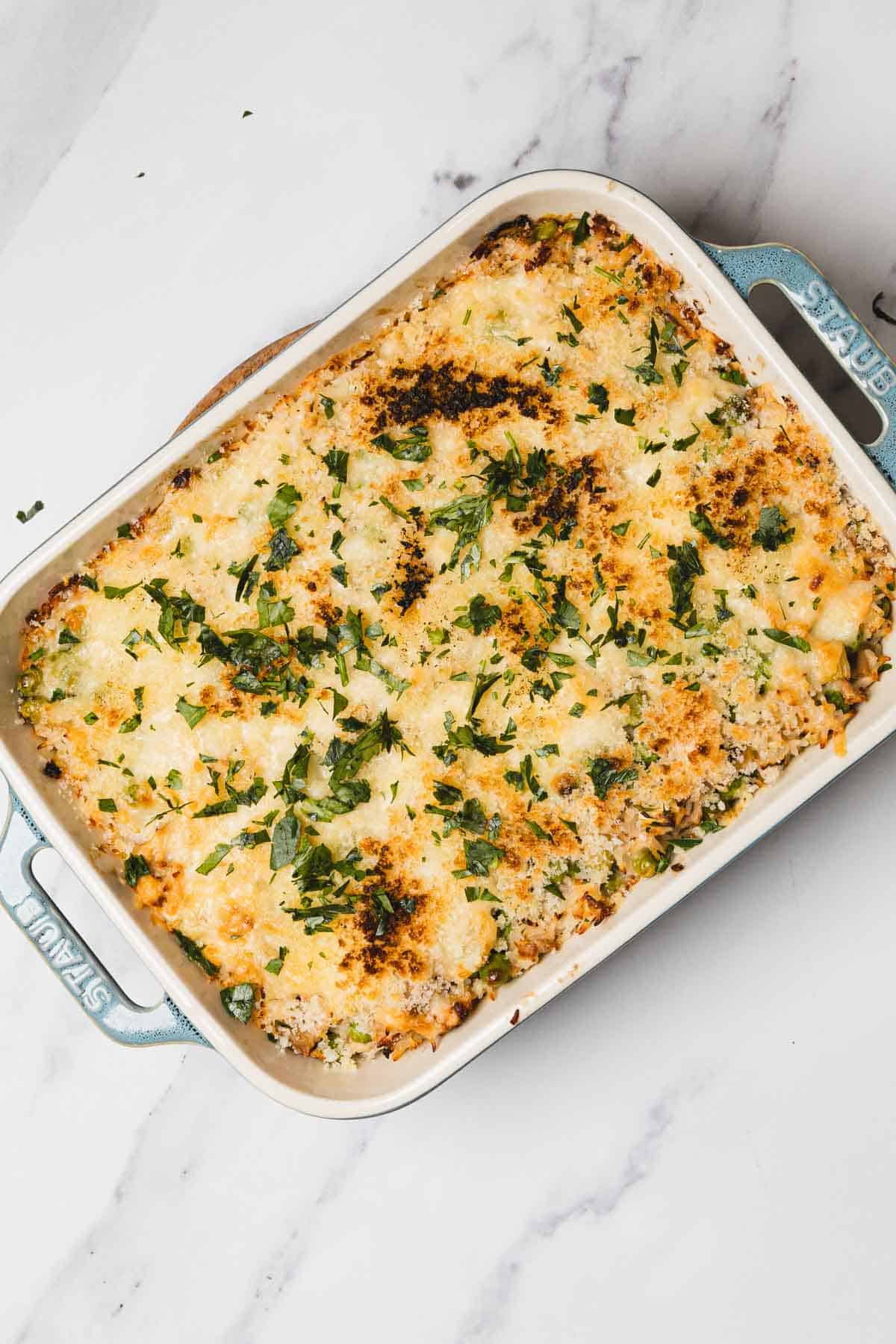 Casserole dish filled with baked cheesy tuna casserole topped with herbs on a marble surface.